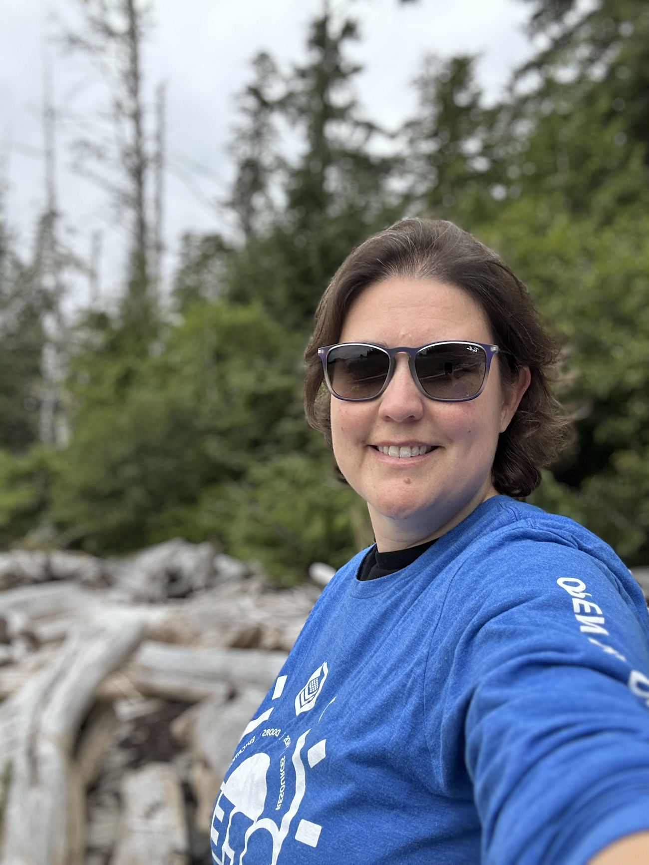 Woman in a blue shirt taking a selfie on a beach with the forest in the background